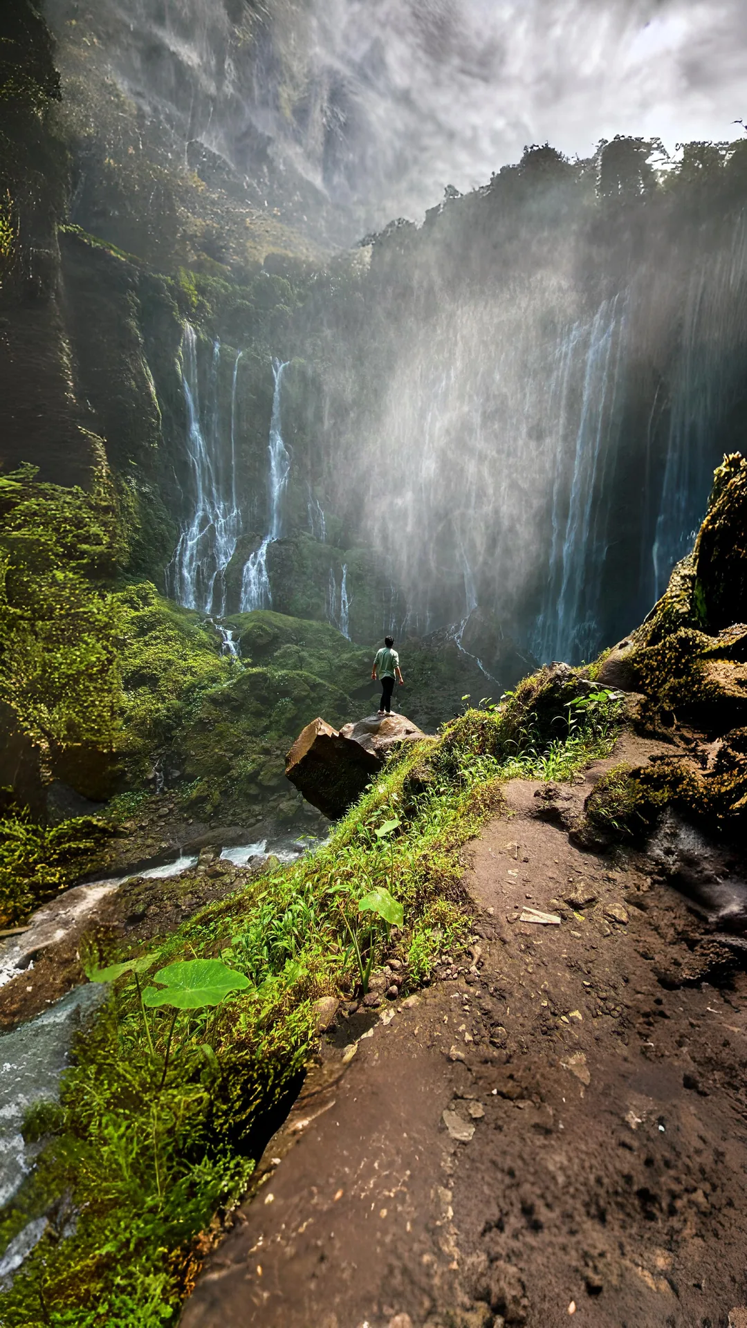 A visitor standing on the rocky path beneath the towering Tumpak Sewu waterfall