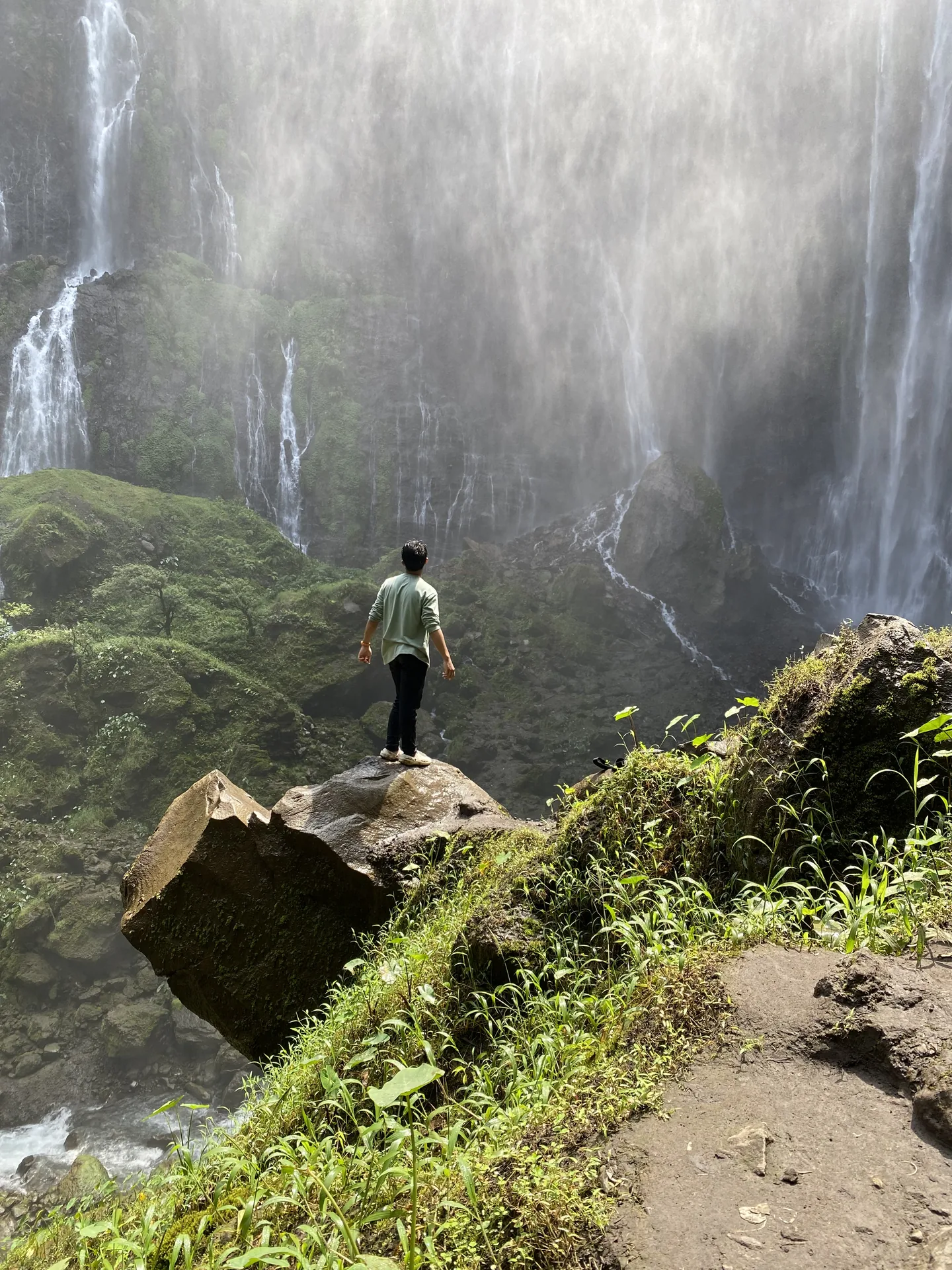A person standing among rocks, greenery, and drifting mist below Tumpak Sewu waterfall